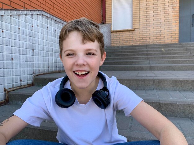 Un niño está sentado en una escalera de hormigón con auriculares y una camiseta blanca sonriendo brillantemente