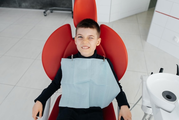 El niño está sentado en una silla dental roja y sonriendo en odontología blanca moderna Tratamiento y prevención de caries desde la infancia Odontología moderna y prótesis