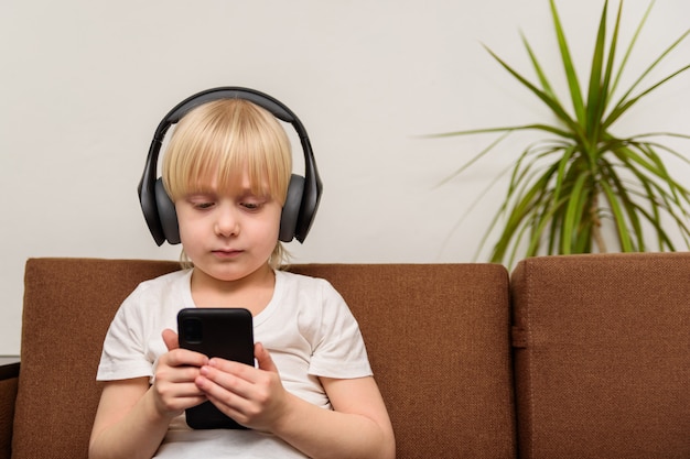 El niño está sentado en el sofá con auriculares y mira fijamente el teléfono. Concepto de adicción a niños y gadgets