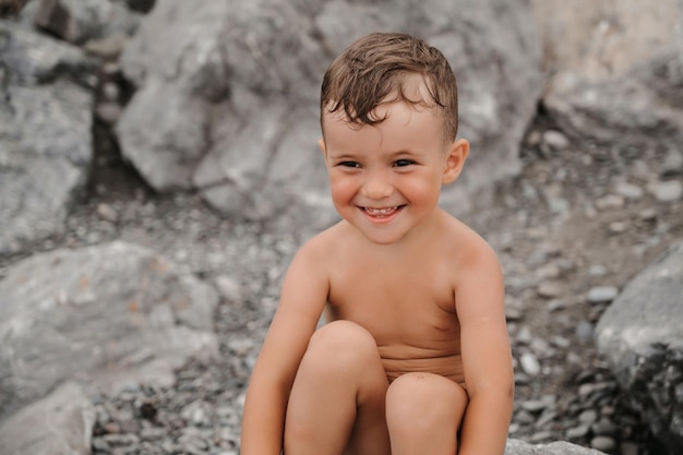El niño está sentado sonriendo y tomando el sol en grandes rocas en la playa cerca del mar