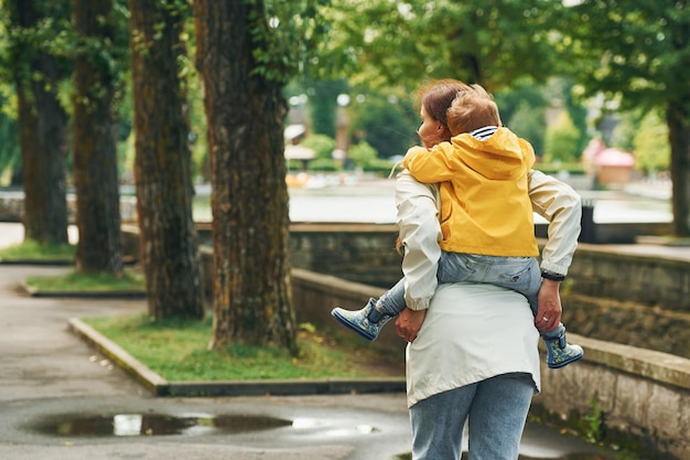 El niño está sobre los hombros La madre con su hijo está dando un paseo al aire libre en el parque después de la lluvia