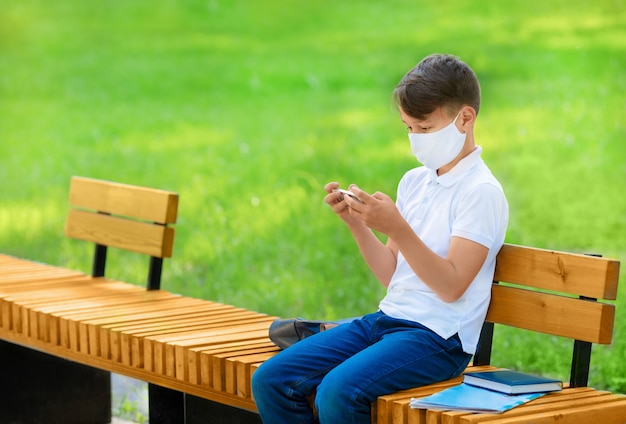 Niño estudiante con máscara está sentado en un banco y sosteniendo el móvil durante la pandemia de covid 19