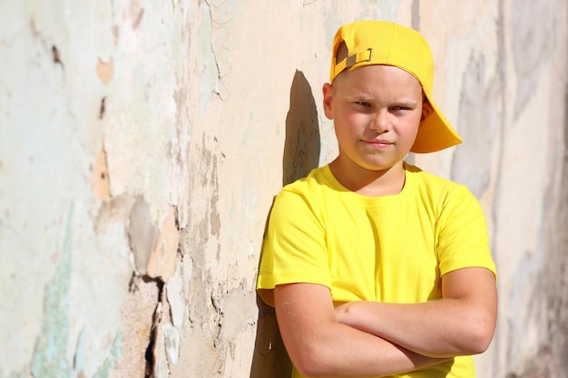 Un niño europeo con una camiseta amarilla y una gorra está de pie contra la pared bajo el sol brillante del verano. Foto de alta calidad
