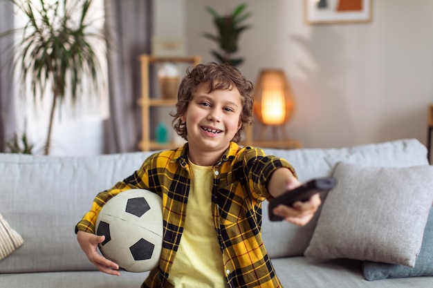 Un niño fanático del fútbol viendo la televisión en casa sentado en un sofá con control remoto y una pelota de fútbol sonriendo a la cámara