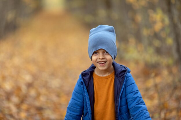 Niño feliz y alegre con ropa de abrigo de colores brillantes jugando entre hojas de arce caídas y recogiendo ramo seco Rayos de sol cayendo a través de los árboles en el parque forestal de otoño al atardecer