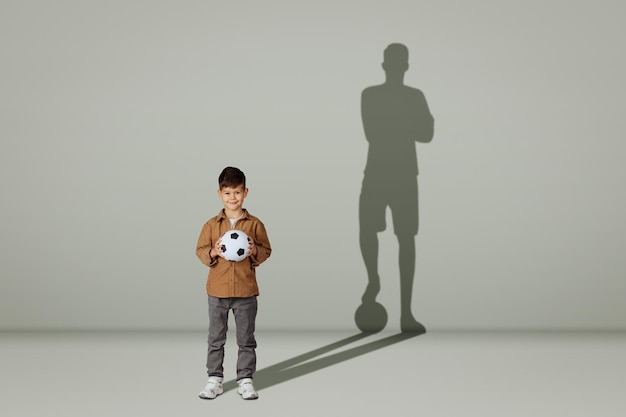 Niño feliz de años caucásicos en casual con balón de fútbol y sombra de jugador de fútbol adulto