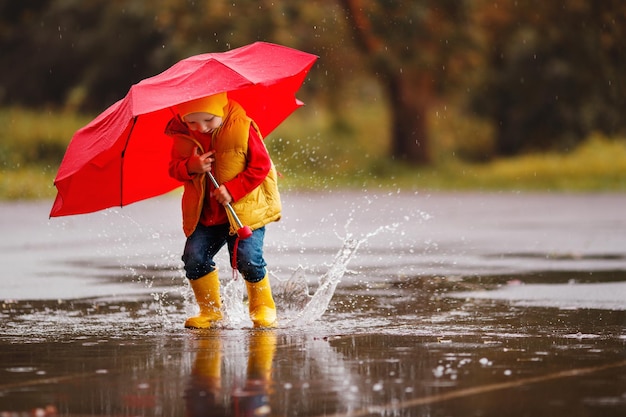 Niño feliz bebé niño con botas de goma y paraguas salta en charco en caminata de otoño
