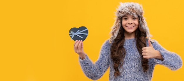 Niño feliz cabello largo con sombrero con caja de regalo que muestra el pulgar hacia arriba sobre fondo amarillo Banner de niña con sombrero de invierno encabezado de póster de estudio con espacio de copia