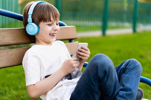 Un niño feliz en la calle se sienta en un banco con auriculares y un teléfono y sonríe