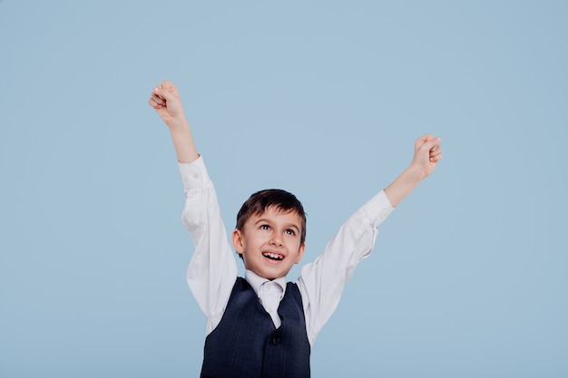 Niño feliz en camisa blanca y chaleco levantando las manos en estudio sobre fondo azul.