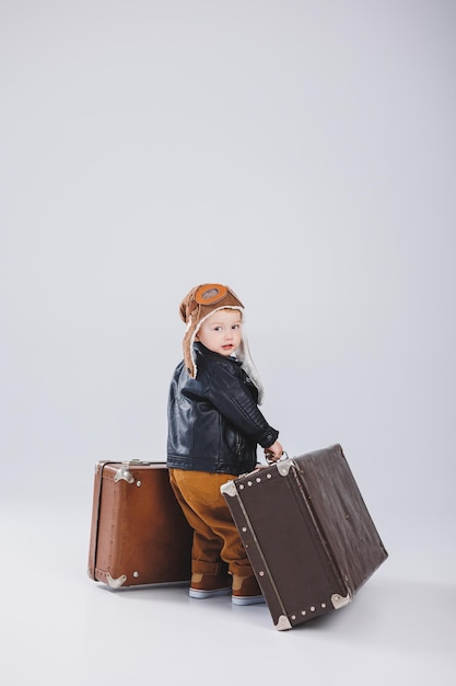 Un niño feliz con casco y una chaqueta de piloto lleva una maleta marrón Retrato de un niño piloto un niño con una chaqueta de cuero Pequeño viajero con una maleta