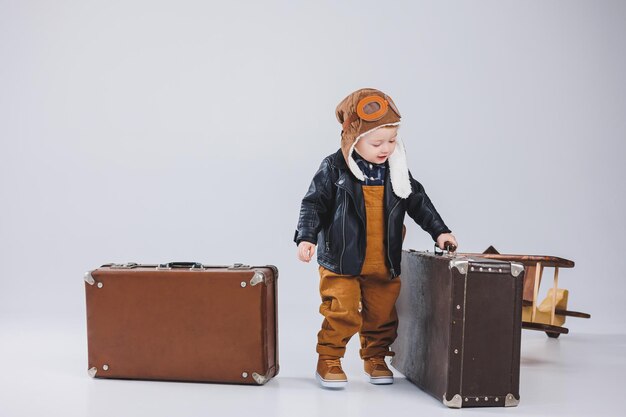 Un niño feliz con casco y una chaqueta de piloto lleva una maleta marrón Retrato de un niño piloto un niño con una chaqueta de cuero Pequeño viajero con una maleta