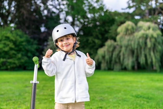 Foto niño feliz con casco de seguridad permanecer cerca de patinete. ciérrese encima del retrato de un niño sonriente que camina en un parque.