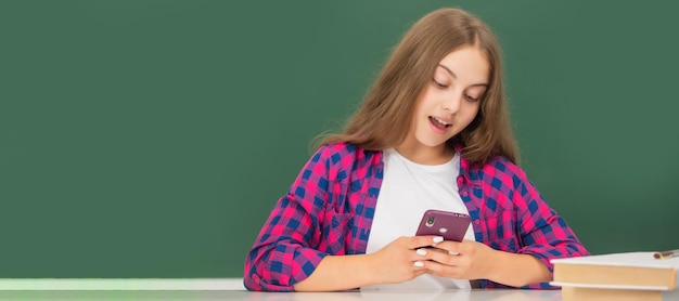 Niño feliz chateando en el teléfono inteligente en el aula en el mensaje de pizarra Cartel aislado horizontal de la estudiante de la escuela Retrato del encabezado de la bandera del espacio de copia de la colegiala