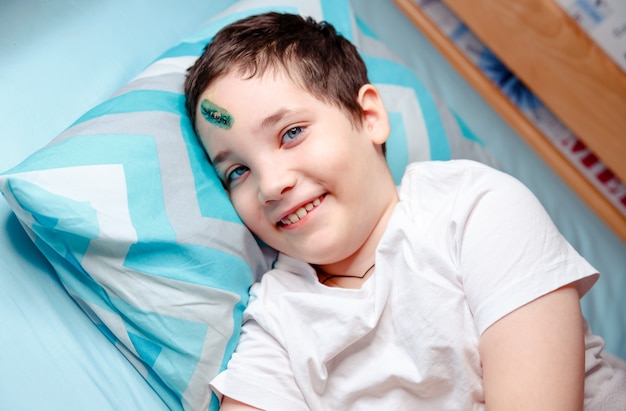 Un niño feliz con una cicatriz en la frente miente y sonríe. El niño está contento porque lo están dando de alta de la habitación del hospital a su casa.