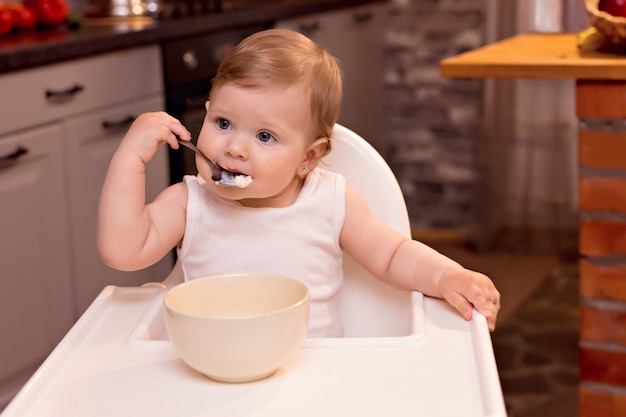 Niño feliz come gachas de leche con una cuchara. Retrato de una niña feliz en una trona en la cocina