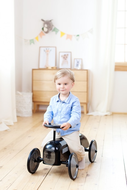 Niño feliz conduce un coche de juguete vintage en una habitación infantil. Niño gracioso jugando en casa. Niño activo conduciendo el coche de un niño en el jardín de infantes. Niño conduciendo un automóvil retro, niño en un automóvil de juguete