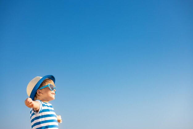 Niño feliz divirtiéndose al aire libre contra el fondo del cielo azul. Retrato de niño sonriente en vacaciones de verano. Concepto de libertad y viaje.