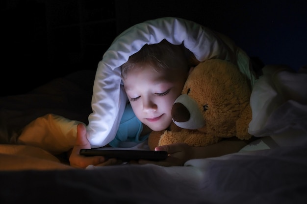 Niño feliz se encuentra con oso de juguete en la cama debajo de una manta y usando un dispositivo de teléfono inteligente tableta digital en la oscuridad. La cara del niño está iluminada por un monitor brillante.