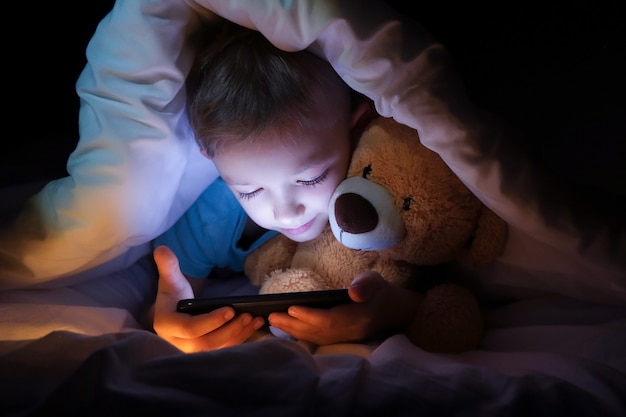 Niño feliz se encuentra con oso de juguete en la cama debajo de una manta y usando un dispositivo de teléfono inteligente tableta digital en la oscuridad
