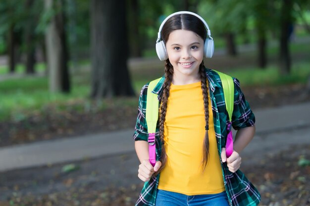 Un niño feliz escucha un curso de audio con auriculares modernos que llevan una mochila escolar en un estilo de moda informal que escucha un paisaje natural de verano