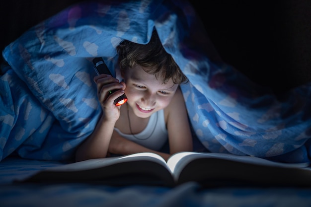 Niño feliz está acostado debajo de la manta y leyendo un libro con una linterna, en la noche