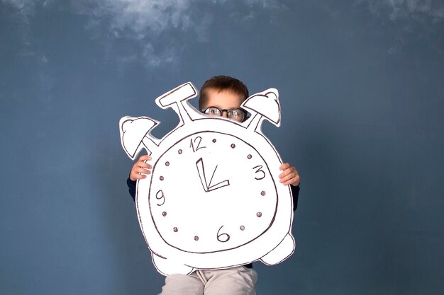 El niño feliz está sentado en un suéter azul con un gran reloj Concepto de modo horario descanso El niño regresa a la escuela Concepto de educación y tiempo