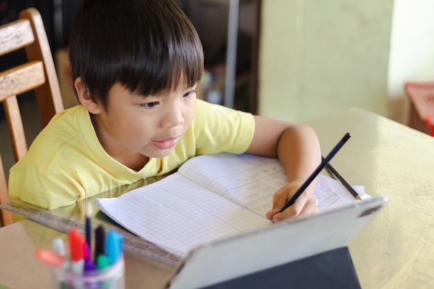 Niño feliz estudiante asiático usando y tocando la almohadilla o tableta inteligente para hacer sus deberes y en línea