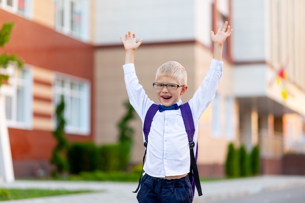 niño feliz con gafas y un maletín está feliz con las manos en alto cerca de la escuela