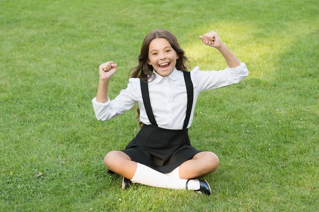 Niño feliz ganador en uniforme escolar muestra el signo ganador de la victoria sentado en la beca de hierba verde al aire libre