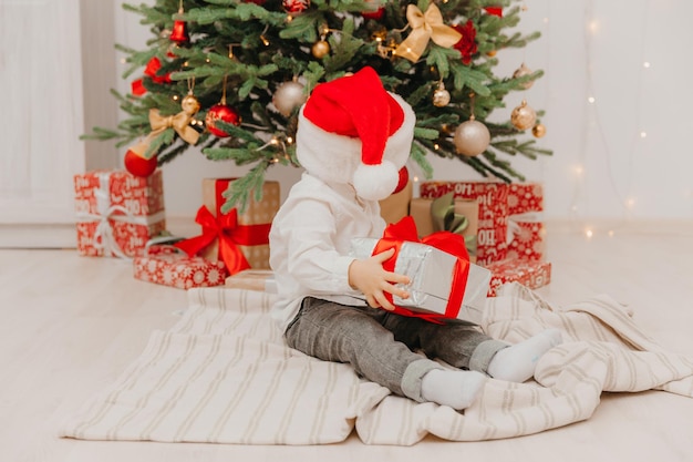 Un niño feliz con un gorro de Papá Noel se sienta en el suelo cerca del árbol de Navidad y sostiene una caja con un regalo en sus manos.