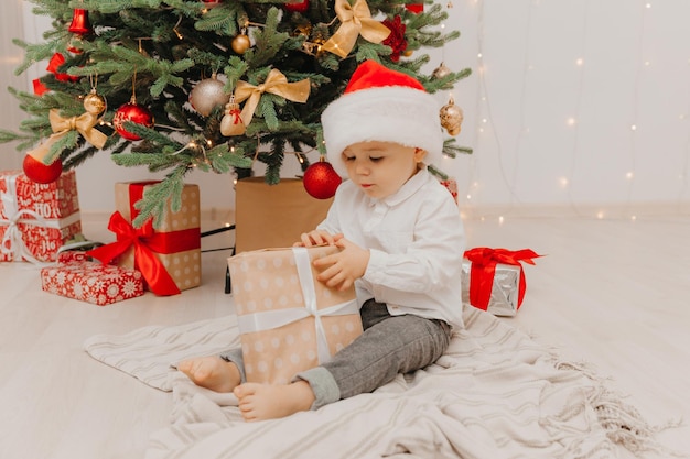 Un niño feliz con un gorro de Papá Noel se sienta en el suelo cerca del árbol de Navidad y sostiene una caja con un regalo en sus manos.