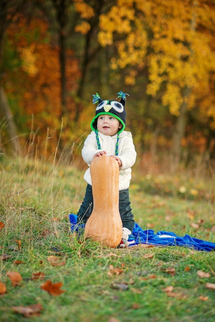 Niño feliz en un gorro de punto y ropa de abrigo con calabaza en el césped entre los árboles de otoño.