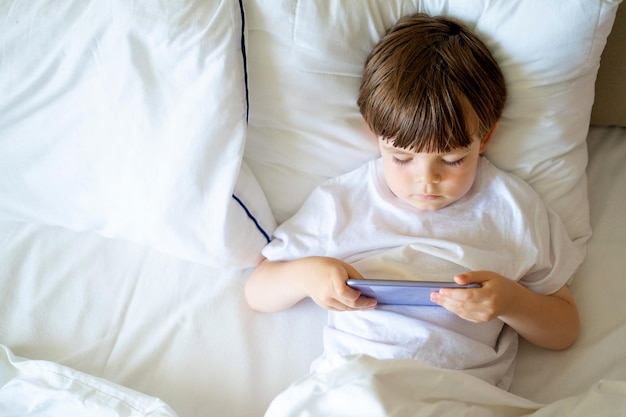 Niño feliz hablando por teléfono inteligente mirando a un lado mientras está sentado en la cama