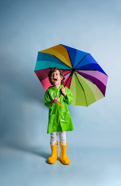 Un niño feliz con un impermeable verde y botas de goma amarillas sostiene un paraguas multicolor sobre un fondo azul con una copia del espacio