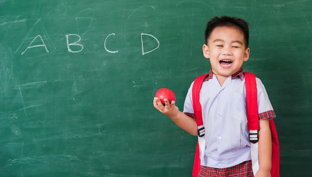 Niño feliz de jardín de infantes en uniforme de estudiante con mochila escolar sostenga manzana roja en la mano