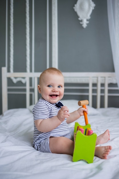 Niño feliz juega con un juguete de madera en una cama blanca y mira a la cámara