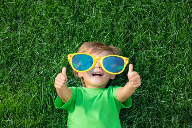 Niño feliz jugando al aire libre en el parque de primavera. Niño acostado sobre la hierba verde. Día de la tierra y concepto de ecología. niño, actuación, pulgares arriba