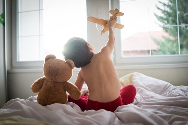 Niño feliz jugando con avión de juguete de madera y oso de peluche en el dormitorio