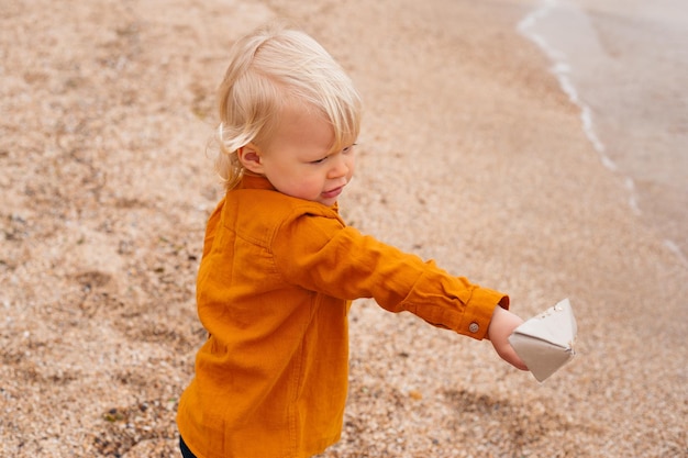 Niño feliz jugando con un barco de papel en la playa en otoño