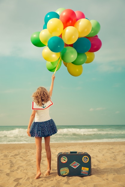 niño feliz jugando con globos de colores y con una maleta en la playa