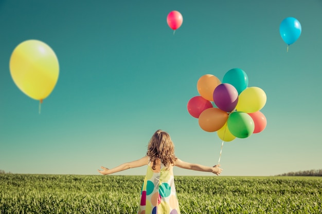 Niño feliz jugando con globos de juguete de colores al aire libre. Niño sonriente divirtiéndose en el campo de primavera verde sobre fondo de cielo azul. Concepto de libertad