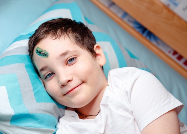 Un niño feliz con una lesión en la cabeza está acostado en la cama y esperando el examen médico en casa.
