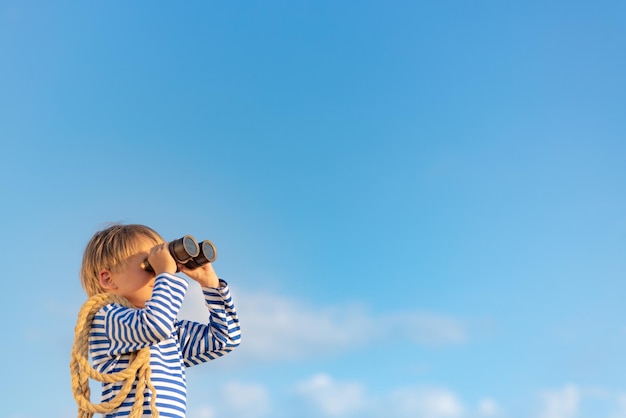 Foto niño feliz mirando a través de binoculares antiguos contra el cielo azul niño divirtiéndose en verano concepto de imaginación y libertad