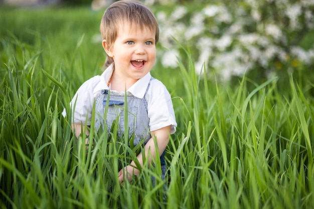 Niño feliz en mono azul de moda con hermosos ojos azules. Juegos divertidos en la hierba verde alta en un parque floreciente lleno de vegetación con el telón de fondo de un manzano.