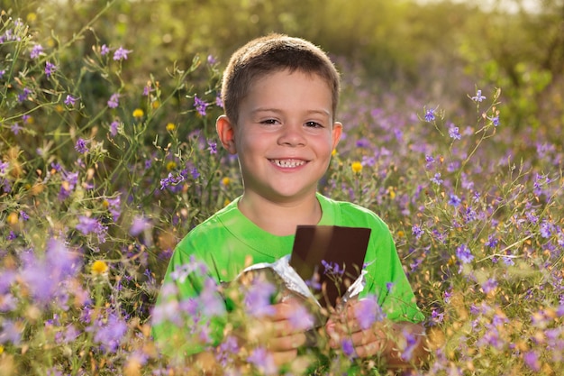 Niño feliz en la naturaleza, con una barra de chocolate en sus manos, rodeado de flores silvestres