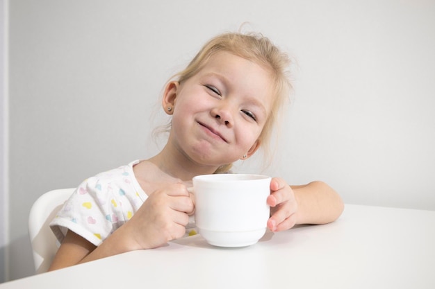 Niño feliz niña rubia bebe de una taza blanca en una mesa blanca