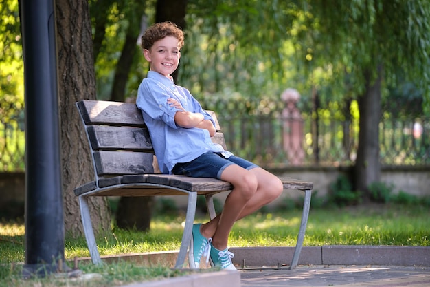 Niño feliz niño relajante sentado en un banco en el parque de verano. Niño positivo disfrutando del verano al aire libre. Concepto de bienestar infantil.