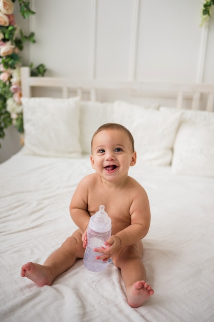 Niño feliz en un pañal sentado en la cama y sosteniendo una botella de agua. La vista desde arriba