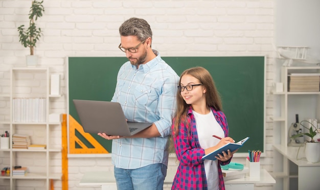 Niño feliz y papá en el aula con cuaderno y computadora en la pizarra, escuela